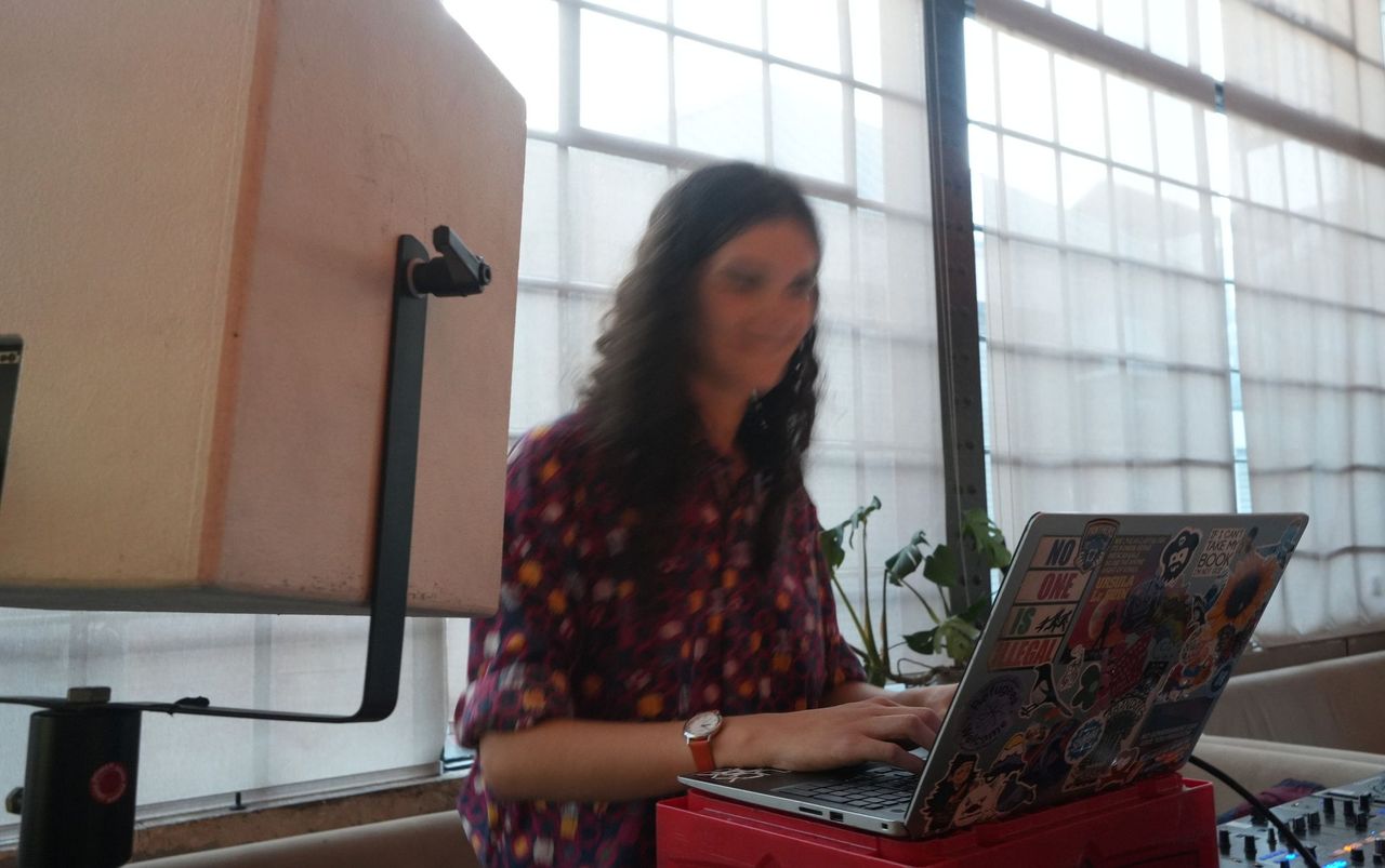 A photo of me playing at an algorave. In the foreground is a DJ monitor and a laptop covered in stickers resting on a bottle crate. In the mid is me, a white person with long brown hair and a bright shirt. My face is blurry (because I'm bopping my head in time with the music). In the background are factory windows covered with linen drapes.