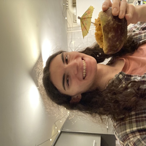A photo of me! A white person with shoulder length, dark brown hair. I'm standing in my kitchen, wearing a red tshirt and chequered shirt. I have a large smile on my face and a turnip filled with lentil stew and a cocktail umbrella in my hand. I look very pleased with myself.